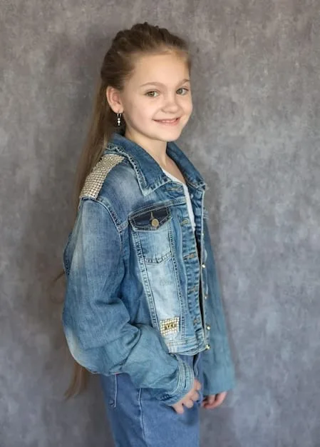 A young girl, around 10 years old, is wearing a denim jacket that she has customized with colorful fabric markers and patches. She's sitting at a table surrounded by art supplies, including markers, patches, and stencils. Her expression is one of concentration and creativity as she works on her design. The background includes other kids engaged in similar DIY fashion projects, creating a lively and artistic atmosphere.