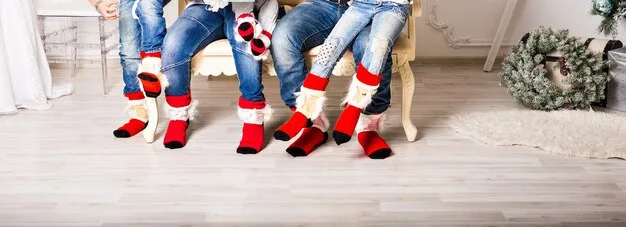 A close-up shot of a family's feet wearing matching Christmas patterned socks, with a blurred background of a decorated Christmas tree. The focus is on the festive sock patterns.
