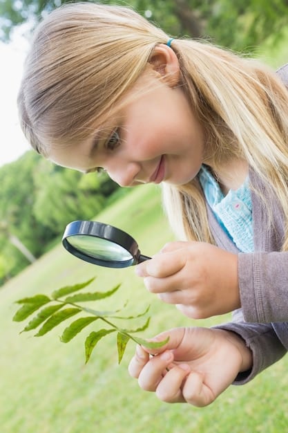 A young girl concentrates as she uses a magnifying glass to examine a leaf during an outdoor science activity with a nature-themed STEM kit.