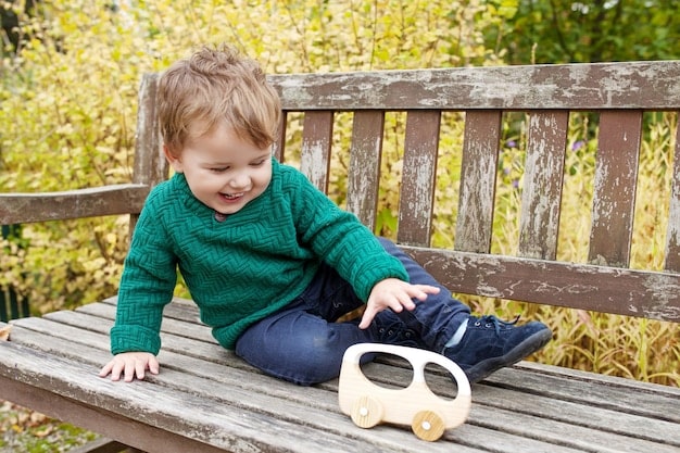 A toddler, approximately 2 years old, happily pushing a small wooden toy car along a smooth surface. The toddler is smiling and engaged, demonstrating the joy of active play.