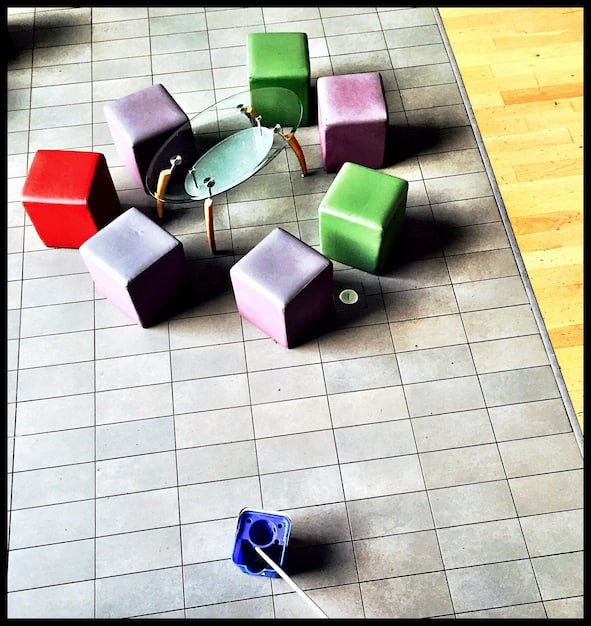 A wide shot of a colorful room floor filled with interconnected magnetic building blocks. The blocks are in various geometric shapes (triangles, squares, pentagons) and bright primary colors. A toddler is kneeling down, reaching for a block, with a curious and engaged expression.