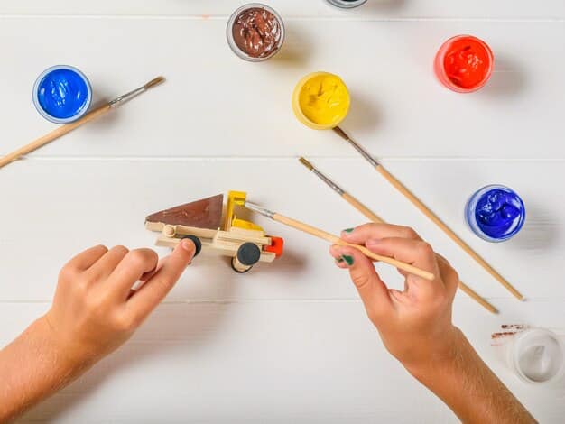 A close-up shot of a child's hands carefully painting a wooden toy car with a small paintbrush. The child is using bright blue paint, and the toy car is partially painted, showing the original wood color underneath. The background is blurred, focusing attention on the painting process and the child's focused expression.