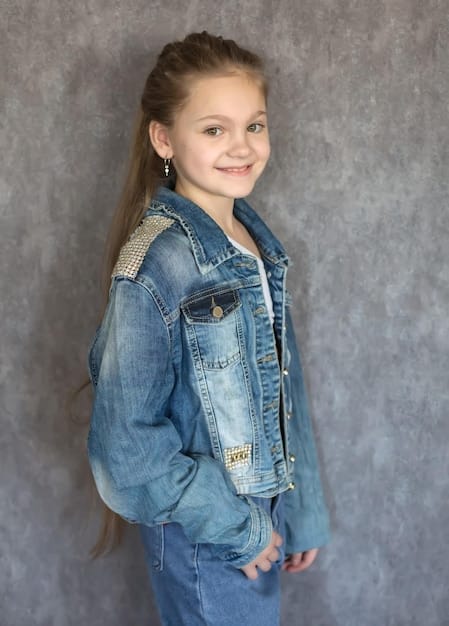 A young girl, around 10 years old, is wearing a denim jacket that she has customized with colorful fabric markers and patches. She's sitting at a table surrounded by art supplies, including markers, patches, and stencils. Her expression is one of concentration and creativity as she works on her design. The background includes other kids engaged in similar DIY fashion projects, creating a lively and artistic atmosphere.